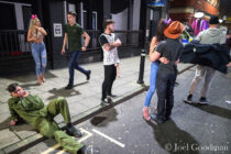 © Joel Goodman - 07973 332324 . 27/12/2016 . Wigan , UK . A man in a Top Gun themed jump suit reclines on the pavement smoking a cigarette next to a man wearing a large prosthetic penis . Revellers in Wigan enjoy Boxing Day drinks and clubbing in Wigan Wallgate . In recent years a tradition has been established in which people go out wearing fancy-dress costumes on Boxing Day night . Photo credit : Joel Goodman